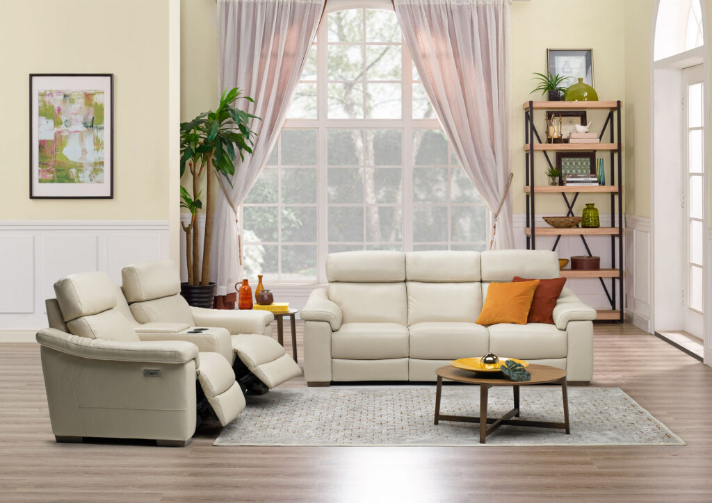 Modern living room with cream leather sofa and recliners, accented by orange pillows. Large window, light curtains, plant, art, and bookshelves create a cozy, airy feel.