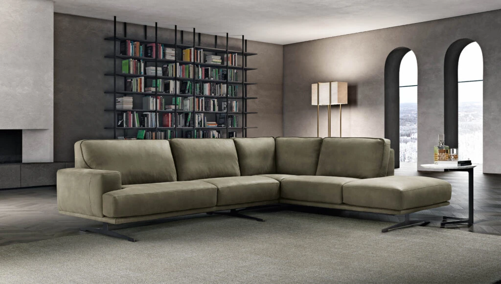 A modern living room with a large beige sectional sofa on a gray rug. A tall bookshelf is filled with books. Two arched windows and a tall lamp add elegance.