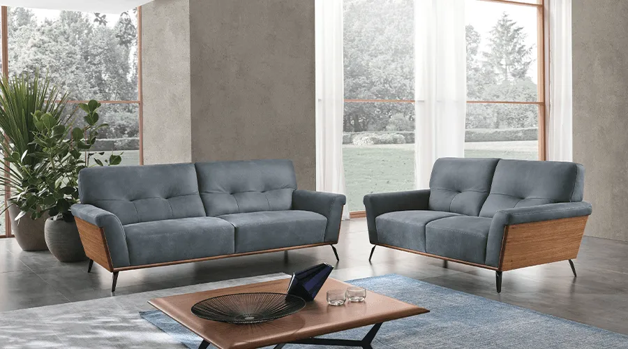 Modern living room with two blue-gray sofas featuring wooden frames, a coffee table, potted plants, large windows, and a serene outdoor view.