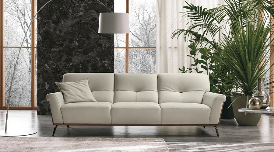 A modern living room features a white sofa with a cushion, framed by large windows showing winter trees. Nearby, lush plants and a standing lamp create a serene, elegant atmosphere.